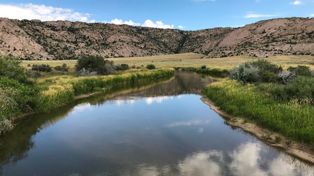 A calm flat river, flat, grassy river banks and rocky bluffs.